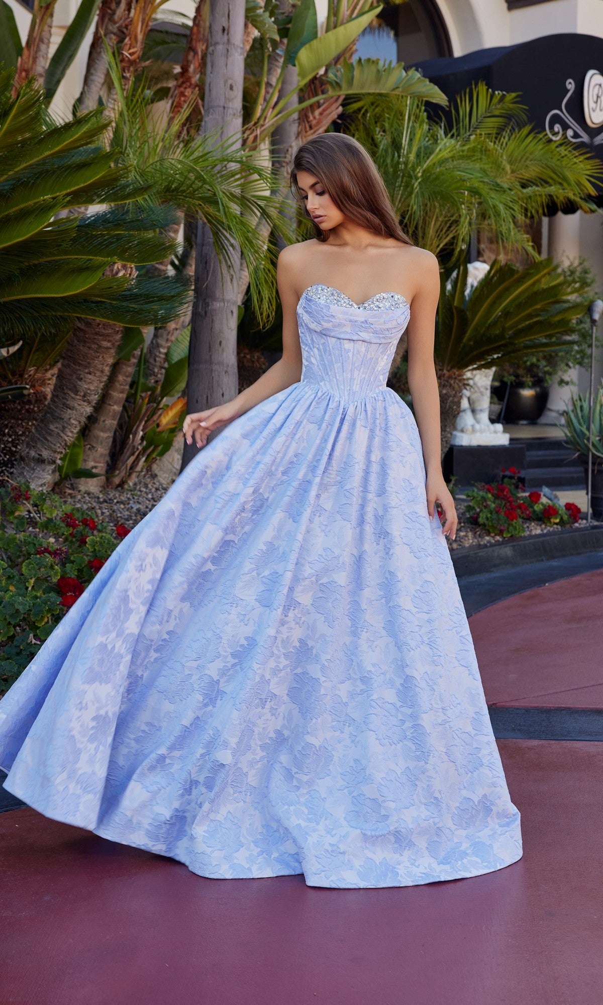 A woman in the Nox Anabel T1912 long prom dress with a jeweled sweetheart neckline stands outdoors near palm trees and red flowers. The lilac, strapless gown shines beautifully in the sunlight.
